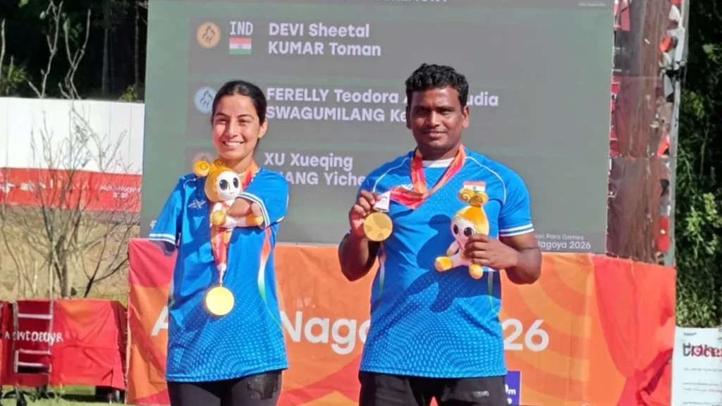 एशियाई पैरा खेलों में नतीजों के बोर्ड के सामने भारतीय किट में कंपाउंड मिक्स्ड टीम के स्वर्ण पदक और खेलों के शुभंकर थामे शीतल देवी और तोमन कुमार