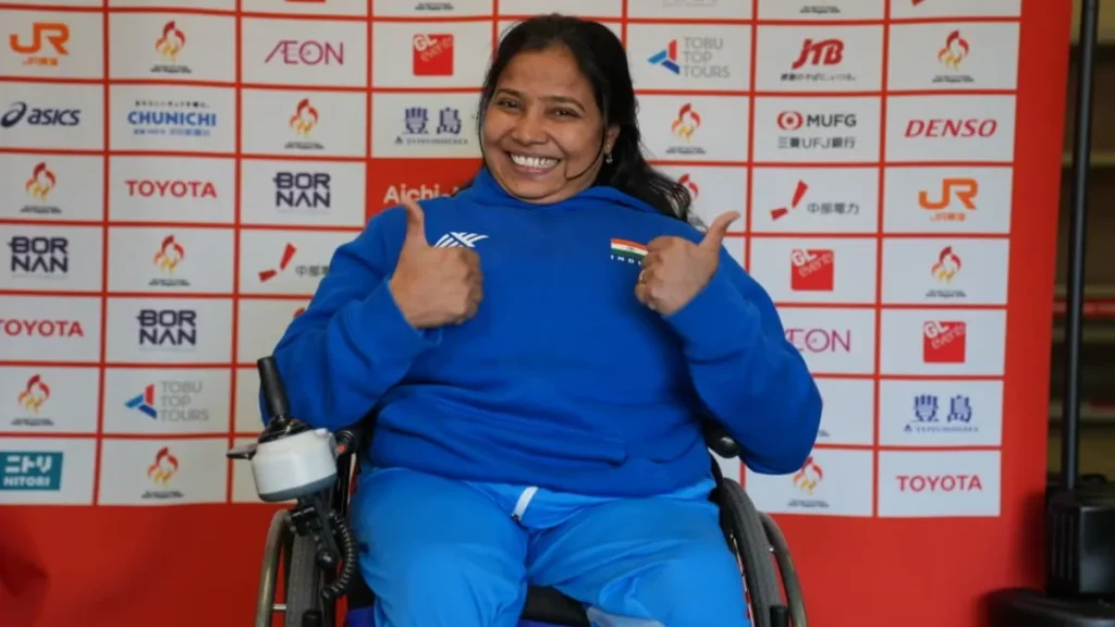 आइची-नागोया 2026 एशियाई पैरा खेलों की प्रायोजक दीवार के सामने नीले भारतीय ट्रैकसूट में व्हीलचेयर से दोनों अंगूठे दिखातीं भारत की सुवर्णा राज